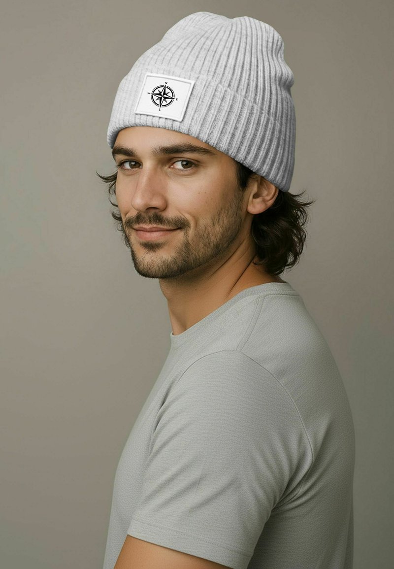 Gray ribbed knit beanie features a rectangular white patch with a black compass design. Model wears a light gray crew-neck shirt.
