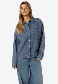 Jeansjacka i mellanblå färg med två bröstfickor, vita knappar, krage och lediga ärmar. Mjuk textur och avslappnad passform.