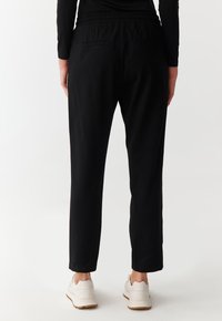 Sorte bukser med elastisk talje, tætsiddende pasform, to baglommer og en blød tekstur. Båret med hvide sneakers, hvilket viser et afslappet look.