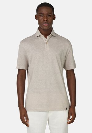 Polo beige à manches courtes, avec un col et une patte de boutonnage à trois boutons, fabriqué à partir d'un tissu doux, porté par-dessus un pantalon de couleur claire.