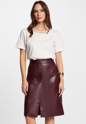 Gonna in pelle bordeaux con taglio dritto, spacco frontale e due tasche applicate; abbinata a una t-shirt bianca con scollatura impreziosita da gemme.
