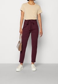 Beige T-Shirt, kurze Ärmel, runder Ausschnitt; bordeauxfarbene Hose mit hoher Taille, Knotenfront, konische Beine; weiße Sneakers; beige Handtasche mit Struktur.
