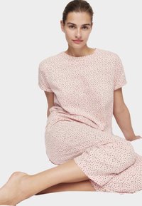 Mujer con el cabello recogido sentada descalza, vestida con una blusa de manga corta, rosa claro con lunares y pantalones a juego, frente a un fondo liso.