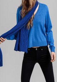 Niebieski sweter z długim rękawem i dekoltem w serek, zestawiony z pasującym, niebieskim dzianym szalikiem o teksturowanym wzorze warkocza. Czarne jeansy.