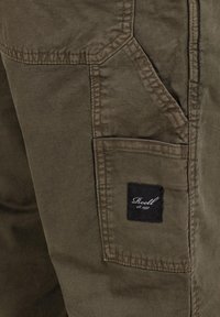 Side pocket of olive-green pants with a black rectangular patch labeled "Reell est. 1997" sewn near the bottom edge.