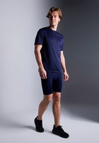 Μπλε ναυτικό αθλητικό μπλουζάκι και εφαρμοστά σορτς από απαλό, ελαστικό ύφασμα, σε συνδυασμό με μαύρα αθλητικά παπούτσια. Το μοντέλο στέκεται μπροστά σε γκρι φόντο.