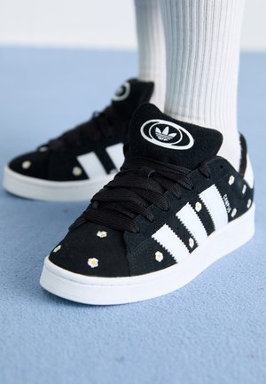 Baskets noires Adidas avec des rayures blanches et de petits motifs de marguerites, portées avec des chaussettes blanches côtelées sur une surface bleue.