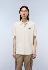 Paita polo berwarna krim dengan lengan pendek, dilengkapi dengan kancing tiga di bagian depan dan logo kecil berwarna hitam di sisi kiri, terbuat dari kain katun.