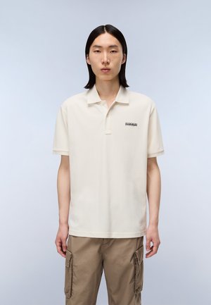 Camiseta polo en color crema con mangas cortas, que presenta una tapeta de tres botones y un pequeño logo negro en el lado izquierdo, fabricada en tela de algodón.
