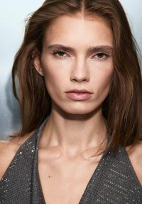 Model met lang bruin haar, gekleed in een grijze halterjurk met glans, heeft een egale huid, gedefinieerde jukbeenderen en minimale make-up.