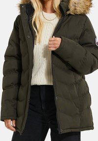 Femme portant une doudoune vert foncé matelassée avec capuche bordée de fourrure, un pull crème en maille torsadée et un pantalon noir, ajustant la fermeture éclair de la veste.