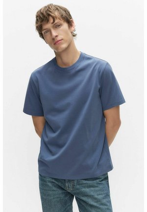 Junger Mann mit hellbraunen Haaren, der ein einfaches blaues T-Shirt und blaue Jeans trägt, steht mit den Händen hinter dem Rücken vor einem weißen Hintergrund.