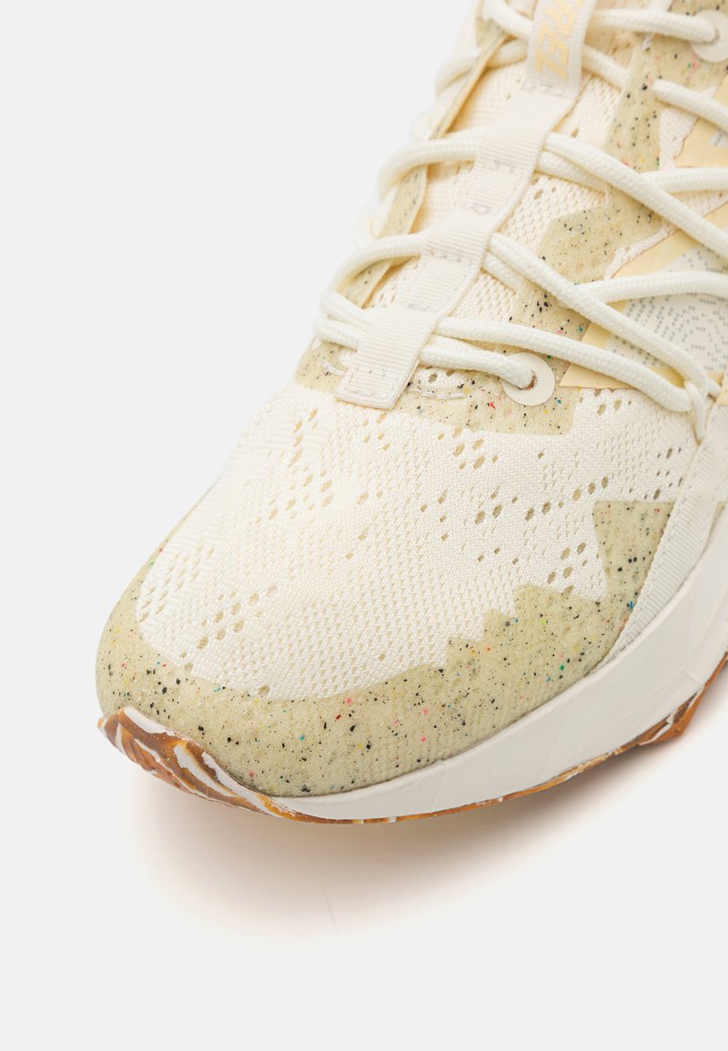 Sneakers blanches avec une tige texturée comportant des perforations, une semelle intermédiaire mouchetée et une semelle extérieure en caoutchouc beige. Comprend des lacets crème et une marque.