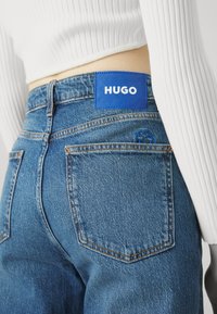 Personne portant un jean en denim bleu avec une étiquette bleue "HUGO" sur la ceinture et un haut blanc côtelé à manches longues.