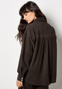 Camisa de pana marrón con un corte relajado, cuello y puños con botones. Presenta un tejido acanalado vertical y un detalle plisado en la parte trasera.