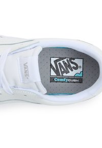 Valkoiset Vans-kengät, joissa on ComfyCush-pohjallinen, ja tasainen ulkopinta, nauhat sekä harmaa sisus, jossa on sinisiä pilkkuja ja logo.