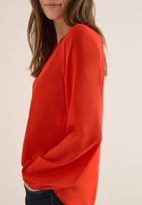 Blusa rossa a maniche lunghe realizzata in tessuto leggero. Presenta un collo a V e una vestibilità morbida, con orlo leggermente curvo e maniche con polsini.