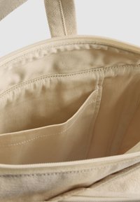 Interior de una bolsa tote de tela beige con una textura suave, que cuenta con cierre de cremallera y un bolsillo lateral para organización.