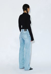 Femme aux cheveux tressés portant un haut noir à manches longues et un jean large taille haute bleu clair, debout de profil sur un fond blanc uni.