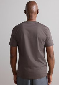 Camiseta de manga corta color marrón, hecha de algodón, con un cuello redondo y una textura suave, mostrada desde la parte de atrás.