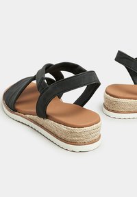 Musta sandaali ristikkäisillä remmeillä, vaaleanruskea pohja, juuttikuidusta valmistettu korko ja valkoinen ulkopohja. Yhtenäinen sileä tekstuuri.