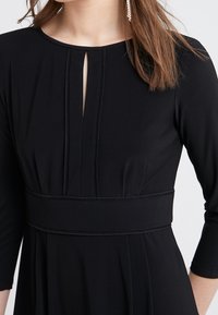 Musta mekko, jossa on pyöreä pääntie avausyksityiskohdalla, kolmanneksen mittaiset hihat ja teksturoitu vyötärö. Sujuva kangas, virtaava siluetti.