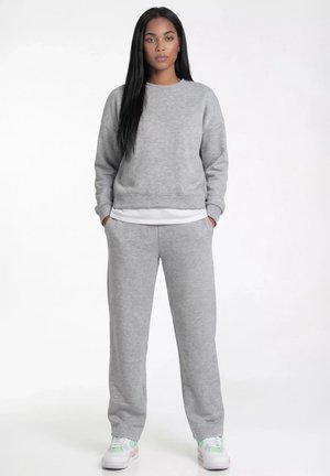 Kobieta stojąca w szarej bluzie i dresach z białymi trampkami, ręce w kieszeniach, neutralny wyraz twarzy, białe tło.