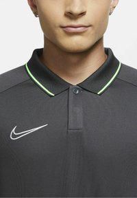 Graues Poloshirt mit knöpfbarem Kragen, mit einem weißen Nike-Logo und einem grünen Akzent am Kragen. Glatte Stoffoberfläche und taillierte Passform.