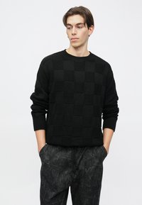 Juodas mezgintas megztinis su tekstūruotu šachmatų lentos raštu, apvalia apykakle ir riešinėmis su raštais, derinamas su tamsiais raštuotais kelnėmis.