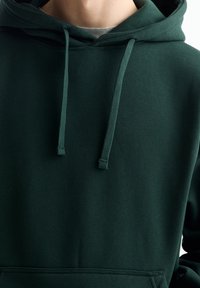 Personne portant un sweat à capuche vert foncé avec des cordons et une poche avant, un t-shirt gris visible sous le col.