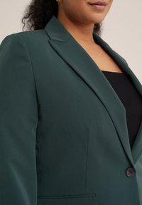 Tmavě zelený blazer s hladkým texturem, se špičatými klopami, zapínáním na jeden knoflík a strukturovaným střihem, nošený přes černý top.