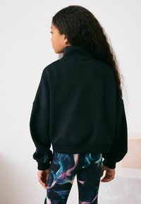 Schwarzer, übergroßer Sweatshirt mit hohem Kragen und herabgesetzten Schultern. Besitzt gerippte Bündchen und einen Bund. Kombiniert mit bunten, gemusterten Leggings.