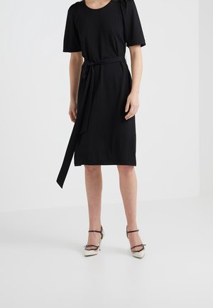 Kobieta w czarnej sukience do kolan z krótkimi bufkami na rękawach, paskiem przewiązanym w talii i białych szpilkach z czarnymi paskami.