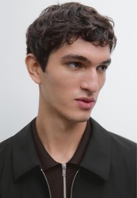 Close-up portrait of a person with short, curly dark hair wearing a black coat over a brown collared shirt, showcasing smooth skin and defined features.