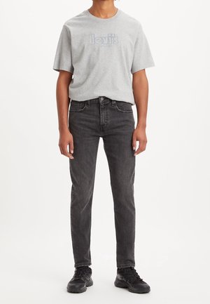 Person trägt ein hellgraues Levi's T-Shirt, schwarze Slim-Fit-Jeans und schwarze Turnschuhe, steht mit den Armen an den Seiten.