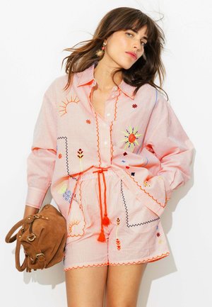 Femme aux cheveux bruns portant une chemise rose à rayures et un short avec des motifs colorés de soleil et de fleurs brodés, tenant un sac en daim marron.