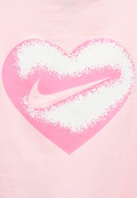 Ροζ καρδιά με λευκό λογότυπο Nike swoosh στο εσωτερικό, σε απαλό ροζ φόντο με υφή σχεδίου.