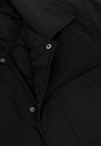 Schwarze gesteppte Jacke mit Druckknopfverschluss, große Klappentasche, strukturiertes Gewebe und gefütterter Innenkragen.