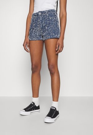 Kvinde iført blå teksturerede strikkede shorts, hvide sokker, sorte lave sneakers og en hvid ærmeløs top, stående mod en ensfarvet væg.