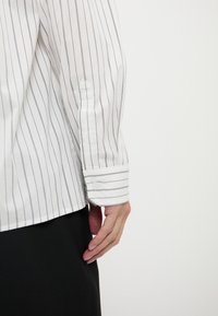 Camisa blanca de manga larga con rayas verticales azul marino; presenta puños abotonados y una textura suave. Mano apoyada sobre pantalón de tela oscura.