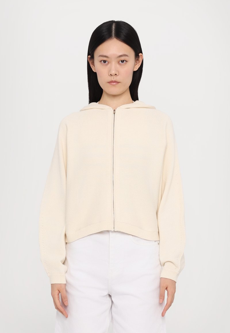 Krémová cropovaná mikina na zips s textúrovaným pleteným vzorom, dlhými rukávmi a dvojdielnou kapucňou. Skombinovaná s bielymi nohavicami.