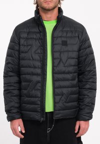 Zwarte pufferjack met gestikte patroon, ritssluiting en hoge kraag. Draagoverlay op een felgroen shirt, voorzien van een logopatch op de borst.