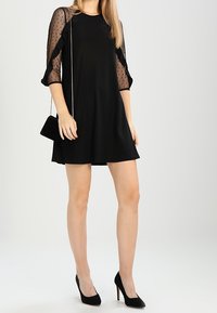 Svart klänning med genomskinliga prickiga ärmar och volangdetaljer, parat med svarta högklackade skor och en liten axelväska.