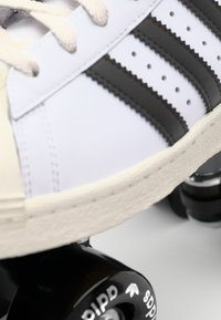 Klassiske hvite sneakers med tre svarte striper, teksturert skinn, rund tå og kremfarget gummisåle, plassert på et svart rulleskatehjul.