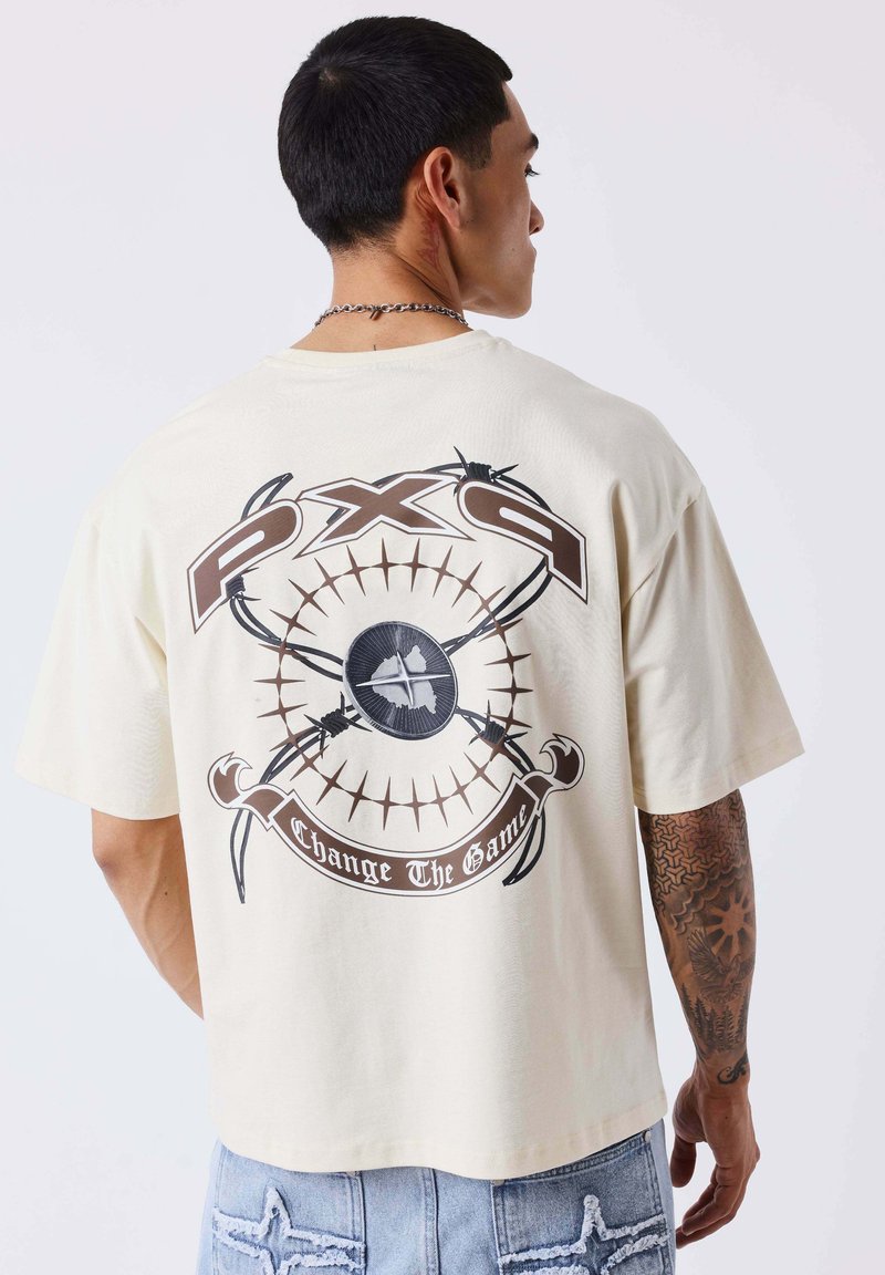 Homme aux cheveux courts noirs et au bras tatoué, portant un t-shirt beige avec un graphisme marron et le texte "PXS Change The Game" dans le dos.