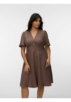 Kvinde med langt mørkt hår iført en brun knælængde kjole med knapper foran og korte ærmer, stående mod en ensfarvet lys baggrund.