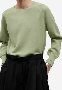 Jasnozielony sweter dziany z prążkowanymi mankietami i okrągłym dekoltem, zestawiony z czarnymi spodniami z wysokim stanem. Prosty wzór o fakturze.