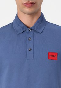 Világoskék poloing, textúrált anyagból készült, bordázott gallérral, három gombbal és egy piros téglalap alakú logóval a bal oldalon.
