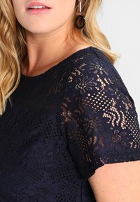 Blusa in pizzo blu navy con motivi floreali e finitura strutturata. Maniche corte con dettagli intricati, creando un effetto a strati.