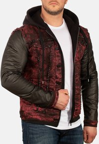 Giacca rossa e nera caratterizzata da un tessuto texture, con cappuccio e maniche in pelle nera, decorata con borchie metalliche lungo il davanti.